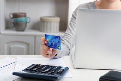 A woman sitting at a table with her laptop and credit card. There is paperwork and a calculator in the forefront.