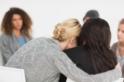 A group of women sitting in a circle having a a group therapy session. Two women in the foreground are sharing a hug.