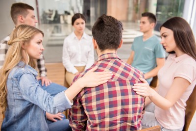 A group of men and women sitting in a circle, Two young girls are comforting a young man.