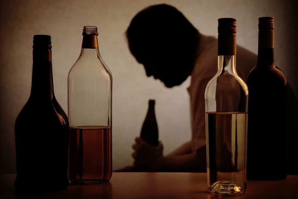 A older male drinking a large bottle of alcohol with 4 other large bottles in the forefront of the image.