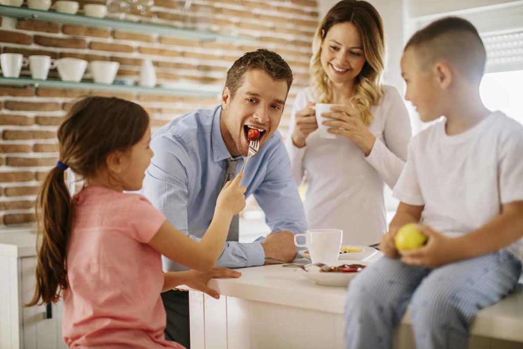 A father being fed strawberries by his daughter at the counter while the mother and son look on smiling.