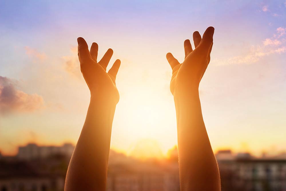 A close up of two hands raised with the setting sun behind them.