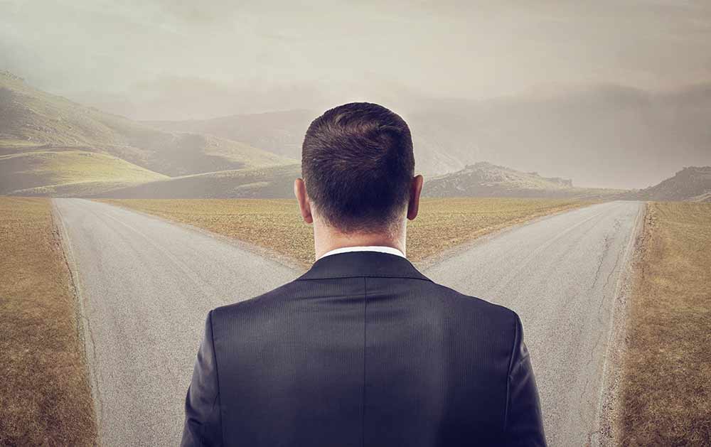 The back of a man's head as he stands before a fork in the road.