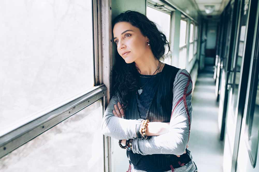 A young woman standing by a window in a long corridor looking out pensively.