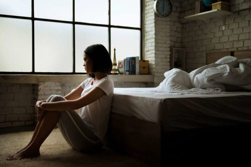 A young woman sitting on floor by an unmade bed looking troubled.