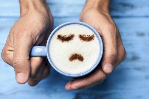 hands holding a mug with sad face in coffee foam