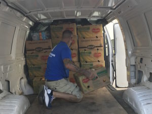 A Beach House volunteer wearing a blue shirt. He is packing a van full of boxes of produce.