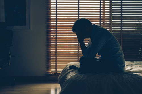 Sad older man sitting with his head in hands on the bed in the dark bedroom.