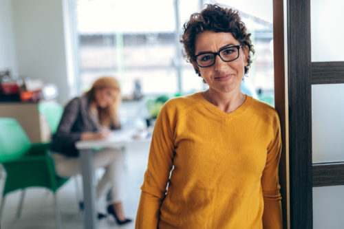 A businesswoman smiling at the camera with another woman sitting at a desk working behind her.