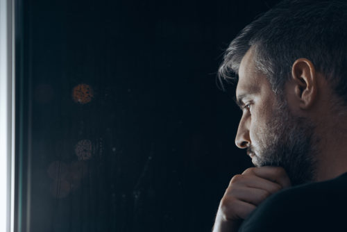 A man with a beard holding his chin beside window at night