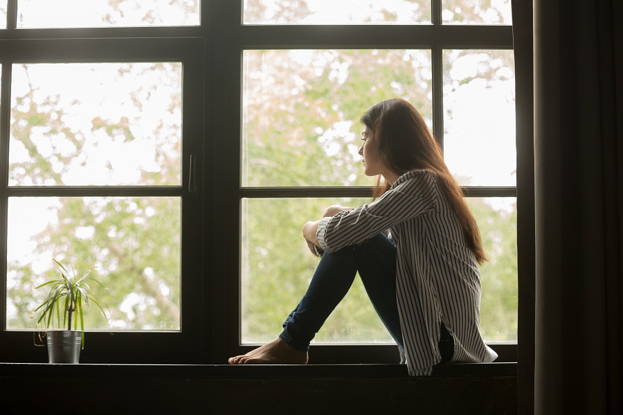 Thoughtful woman sitting on a window sill embracing her knees looking at window