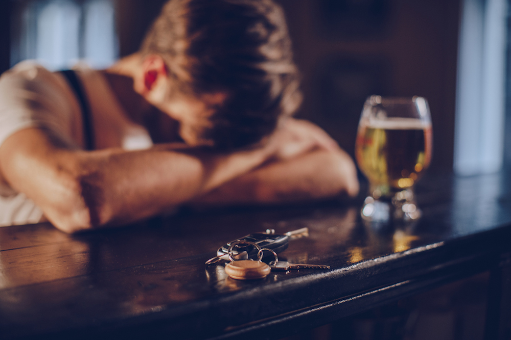 A young man passed out on the bar with a beer in front of him, his car keys are in the forefront of the image.