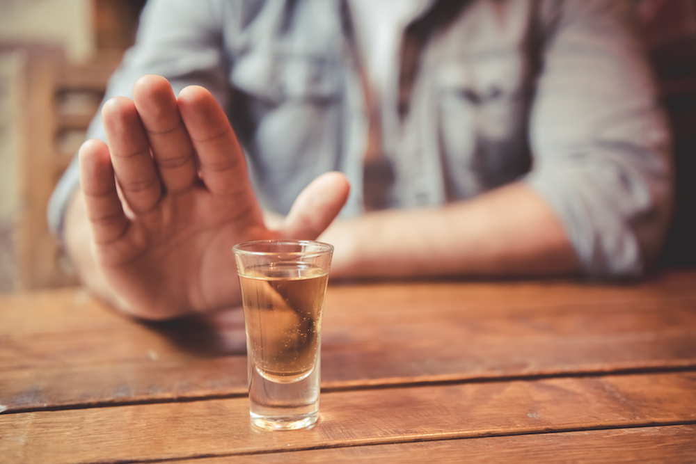 A young man sitting at a table rejecting the shot offered to him.A shot glass of amber liquid is in the forefront.