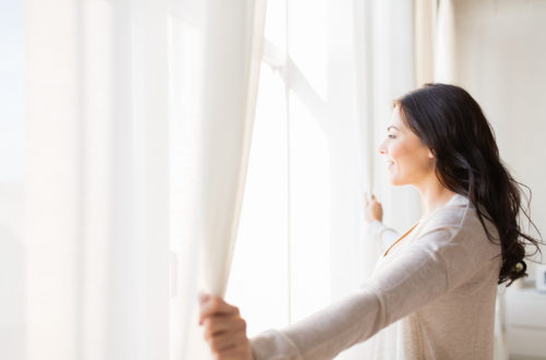 A woman smiling as she opens her curtains on a sunny morning.