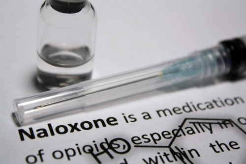 A close up of a needle and medicine bottle laying on a piece of paper with details of the drug Naxolone.
