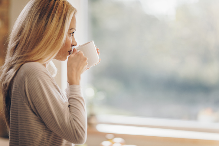 A young woman drinking coffee while looking out the window pensively.