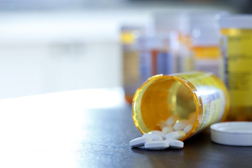A bottle of prescription pills falling out of a bottle with a group of other prescription bottles in the background.