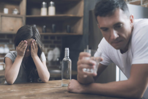 A father and daughter sitting at the kitchen table. the father is drinking a glass of alcohol while the daughter sits with her head in her hands.