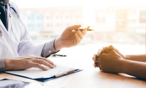 A male doctor filling out paperwork at his desk while talking to a patient.