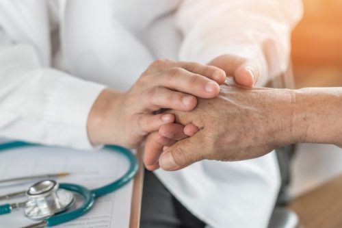 A doctor holding the hand of his patient at his desk. A blue stethoscope lays next to him.