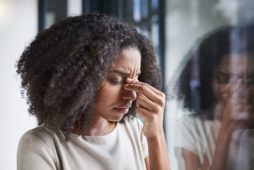 A young woman standing at a window, holding the bridge of her nose while looking agitated.