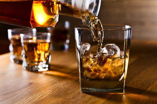 A close up of a glass of whisky being poured into a tumbler full of ice, with two other full glasses in the background.