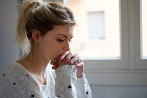 A young blonde woman sitting with her hands by her mouth while looking worried.