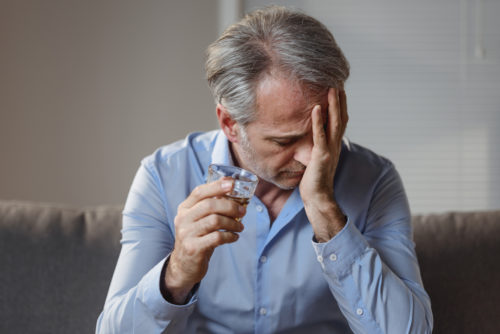 A older male sitting on a couch with his head in his hand and a glass of alcohol in the other hand.