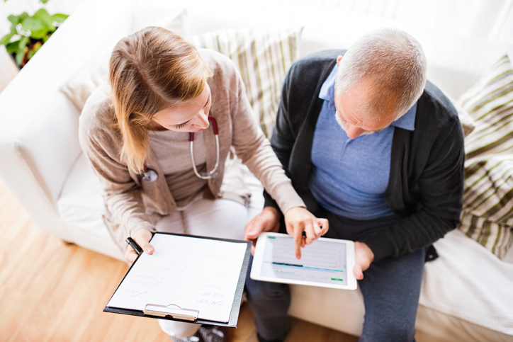 A female health visitor and a senior male. They are sitting on a couch discussing some information on a tablet.