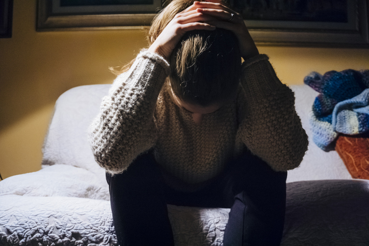 A young woman sitting on the edge of her bed with her head in her hands.