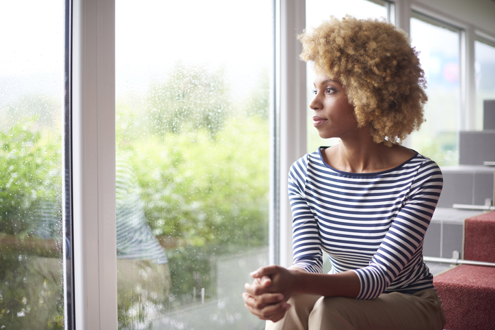 A young woman sitting on some stairs while looking thoughtfully out a window.