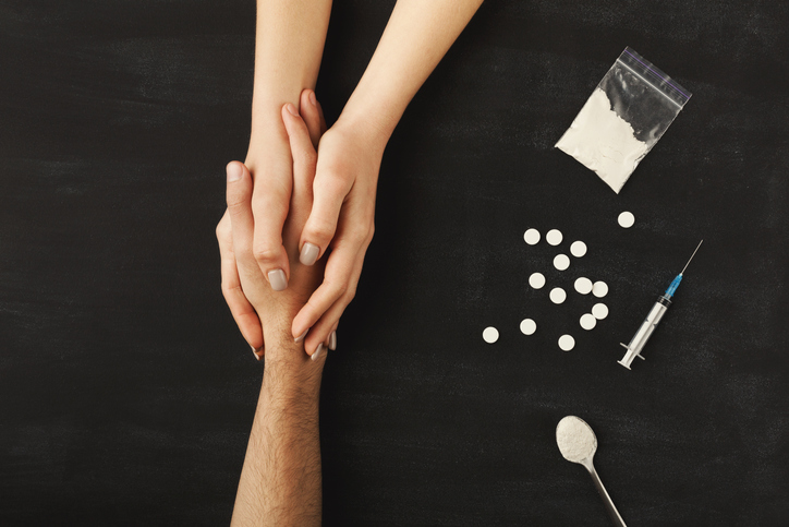A close up of a man and women holding hands with various drugs and drug paraphernalia around them.