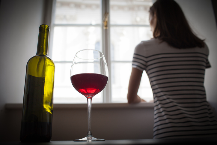 A bottle and glass of red wine on a table. A Young woman stands in the background looking out the window.
