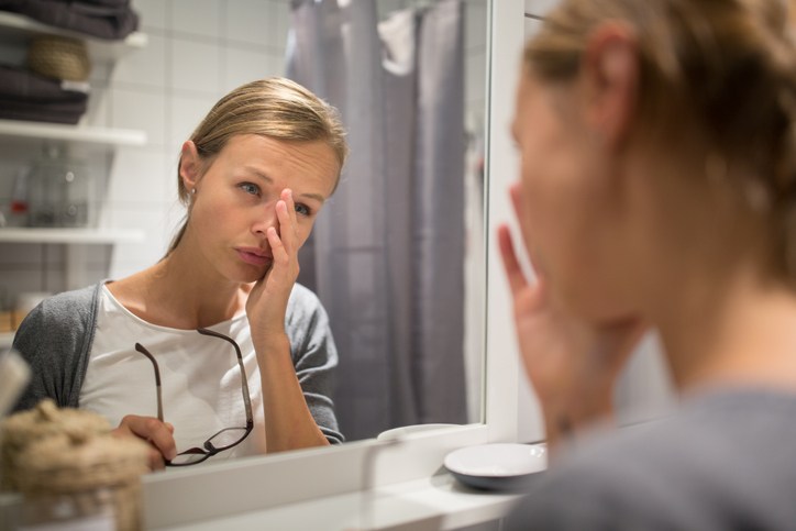 A young woman rubbing her eye in front of her bathroom mirror while holding her glasses.