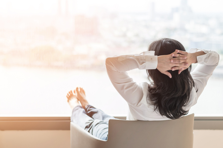The back of a woman who is lounging in a chair with her feet up and hands behind her head.