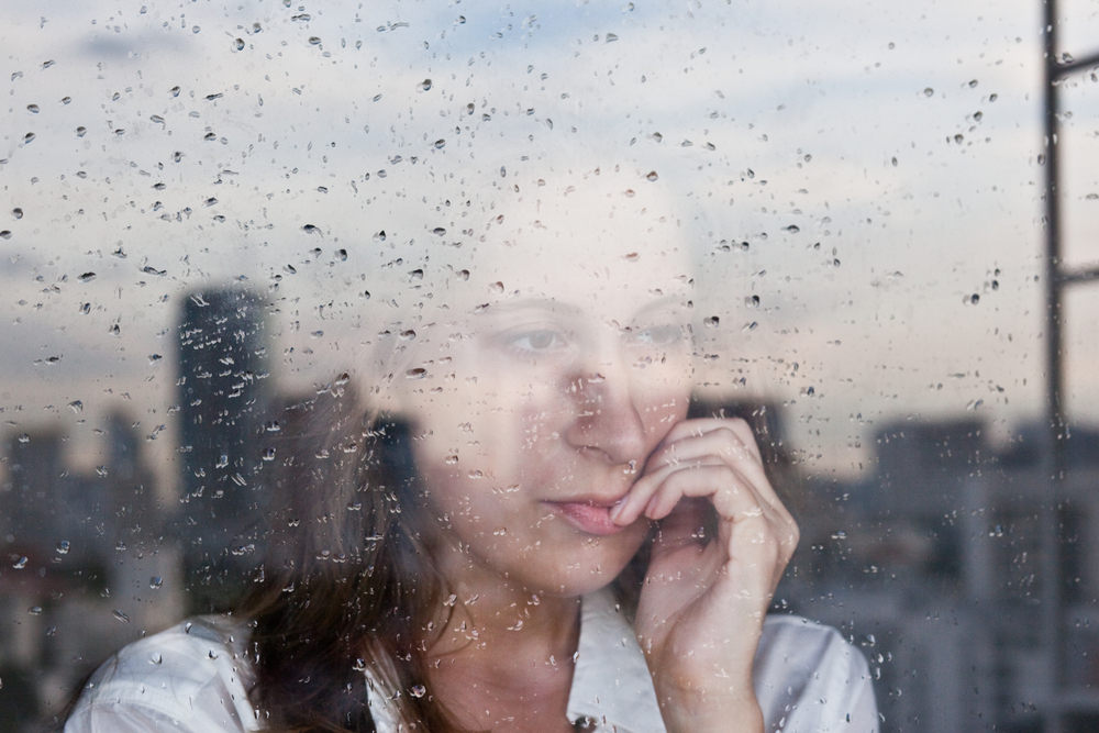 A woman staring out a window pensively with the city reflected in it.