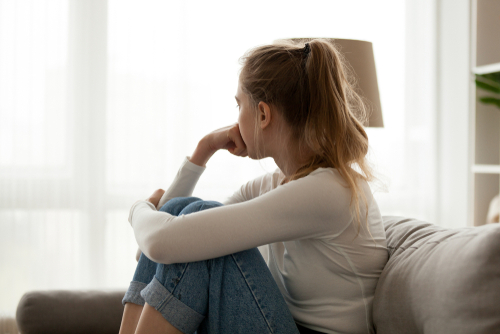 A caucasian woman sitting on a couch looking opensively out a window. She appears troubled.