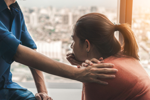 A young woman being comforted by a man as they sit together by a window.