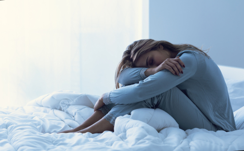 A young woman sitting in bed with her head in her lap, she is in pain or depressed.