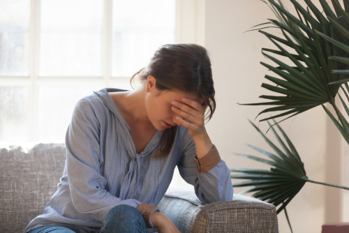 A young caucasian woman sitting on the couch with her head in her hands looking troubled.