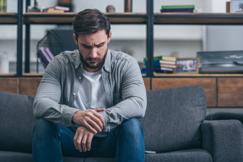 A depressed man sitting and grieving on a couch in his living room.