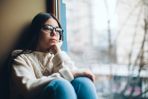 A teenage girl with large glasses seated by a window looking out sadly.