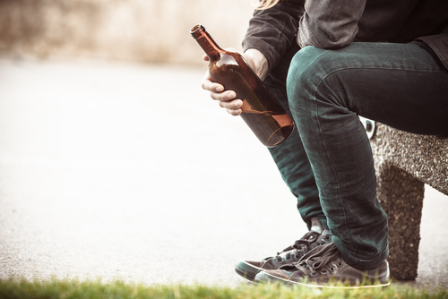 A man depressed with wine bottle sitting on bench outdoor.