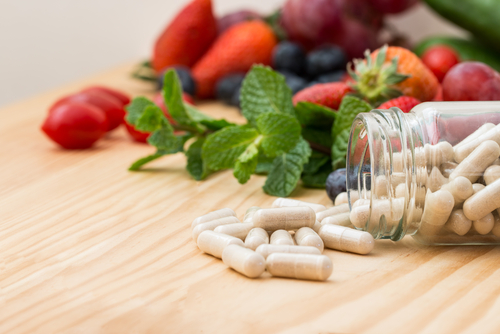Vitamins supplements spilling out of a bottle on wooden table with fruit in the background.