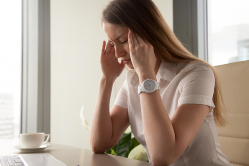 A young woman sitting at a table with her head in her hands, she is in pain with a headache.