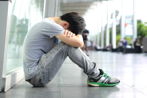 A teenage boy sitting on the floor outside with his head in his lap.
