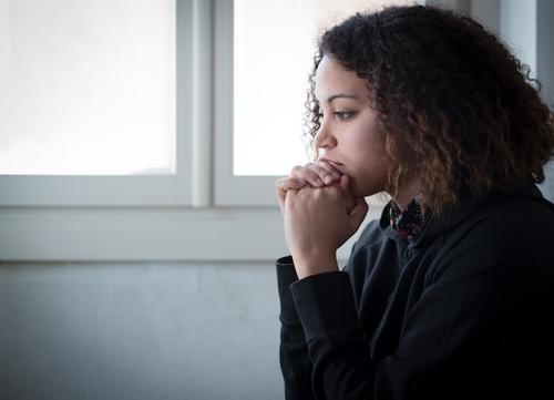 The profile of a young woman sitting near a window looking down pensively.