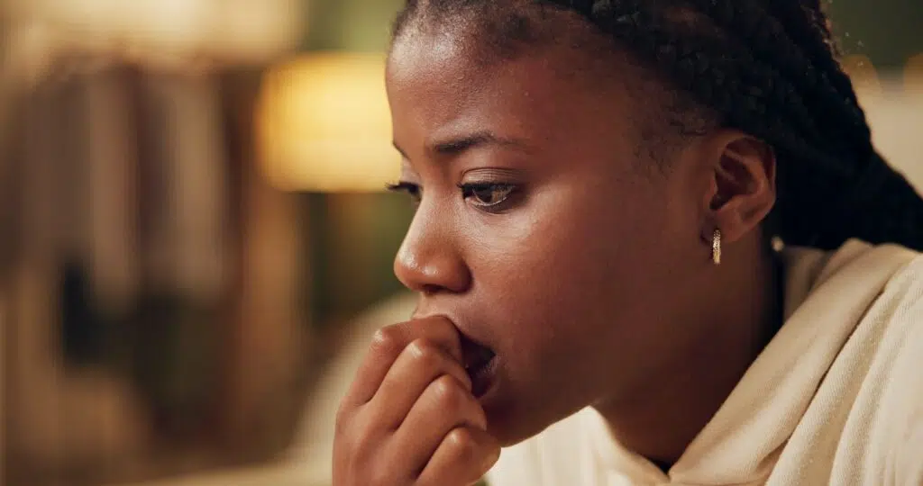 A young Black woman sitting at home, appearing anxious and lost in thought, illustrating stress and worry.