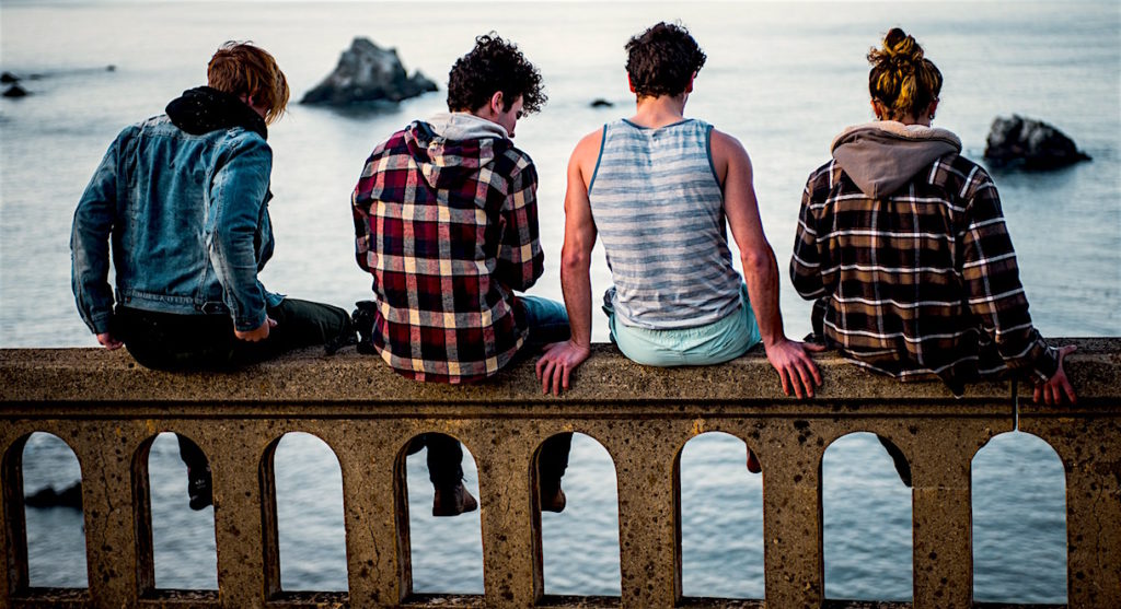 Four caucasian teens sitting on the wall of a bridge, with the ocean below them.