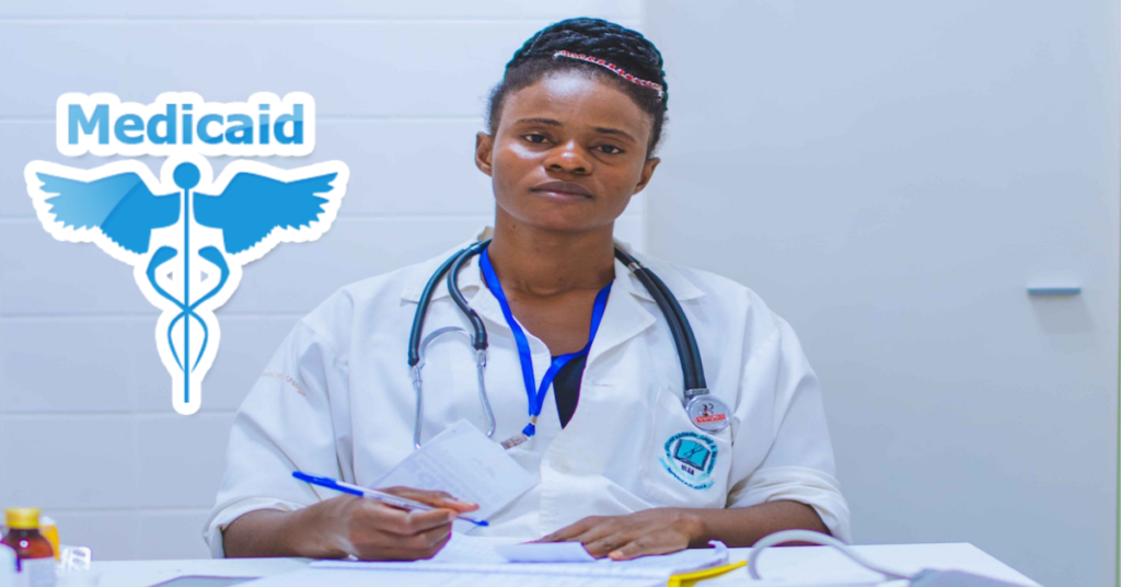 An african-american doctor sitting at her desk filling in paperwork with the medicaid logo on the left of her.